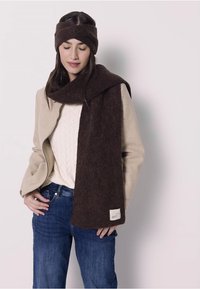 Brun ullscarf med en mjuk textur, bärs draperad över en beige kappa. Kappan har en enkel design och scarfen har en etikettdetalj.