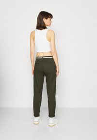 Pantalon vert fuselé avec des ourlets retroussés, des détails de ceinture blanche, et un haut court blanc. Le modèle porte des baskets blanches, tourné le dos à l'appareil photo.