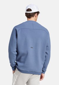 Felpa blu con finitura texturizzata, polsini e orlo a coste. Presenta un piccolo logo sul retro. Abbinata a pantaloni grigi chiari.