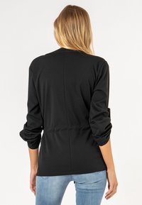 Femme aux longs cheveux blonds portant un haut noir à manches longues froncées et un jean bleu clair, debout devant un fond blanc uni.