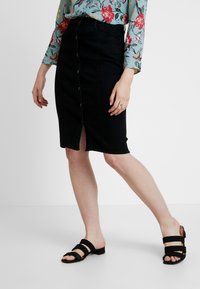 Zwart denim kokerrok met knoopsluiting aan de voorkant, knielang, met een voorkeurspleet. Gecombineerd met zwarte open hak slidesandalen.