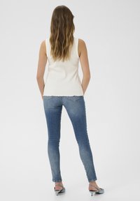 Biely rebrovaný top s tankovým strihom v kombinácii s modrými úzkymi džínami s roztrhnutým lemom, z pohľadu zozadu. Outfit dopĺňajú svetlé lodičky na podpätku.