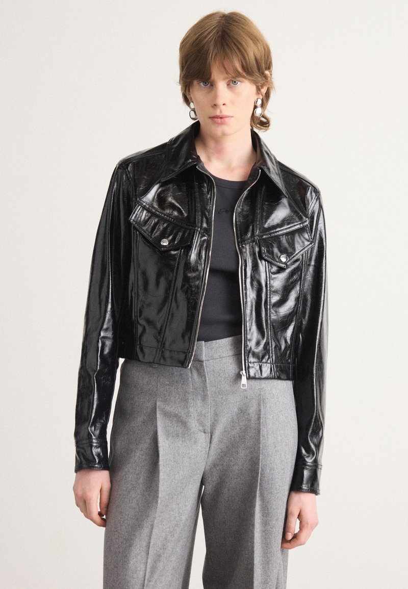 Mustmust nahkade viimistlusega lühike jakk, millel on krae, kaks rinna taskut ja lukuga sulgemine, kombineeritud hallide volditud pükstega.