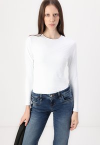 Long-sleeve white top in a smooth, stretchy fabric, paired with blue denim jeans featuring front pockets and a button closure.