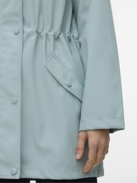 Lähikuva vaaleansinisestä takista, jossa on nepparit, joustava vyötärö, läppätasku ja henkilön käsi lepää rinnalla.