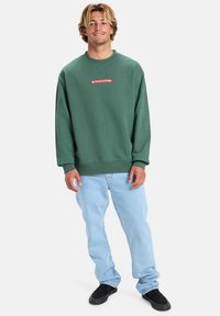 Zelená mikina vyrobená ze směsi bavlny s žebrovanými manžetami, na hrudi s červenou nášivkou loga. Spárována s světle modrými džíny a černými botami.