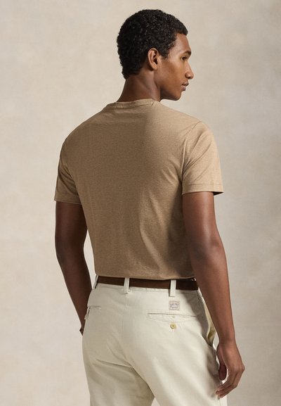 Trumpomis rankovėmis, apvalia iškirpte pasiūtas šviesiai rudos spalvos marškinėliai, aptempto stiliaus, dėvimi su kreminės spalvos kelnėmis su diržu ir subtilia kišenės detalė nugarinėje pusėje.