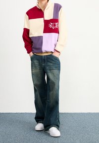 Farveblokeret fleece-pullover i rød, beige, lilla og lavendel. Har en halv lynlås krave og broderet "HUF" logo. Bæres med løse denim jeans.