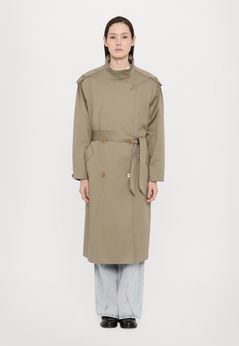 Olive green trench coat with a high collar, double buttons, a belt at the waist, and two side pockets, paired with wide-leg light jeans.