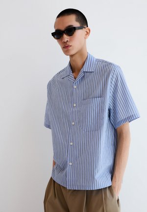 Junger Mann trägt schwarze Sonnenbrille, blaues kurzärmeliges Hemd mit vertikalen Streifen und braune Hose, steht vor einem einfarbigen weißen Hintergrund.