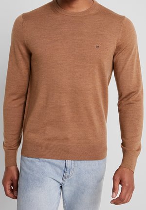 Mężczyzna ubrany w dopasowany, camelowy brązowy sweter z dekoltem typu crewneck z małym logo "ck" na piersi oraz jasnoniebieskie dżinsy.