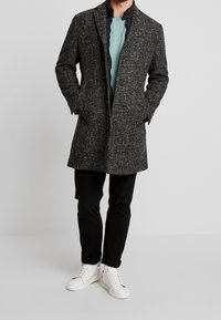 Manteau texturé gris avec une doublure intérieure noire, présentant une coupe ajustée et deux poches avant, associé à un pantalon noir et des baskets blanches.