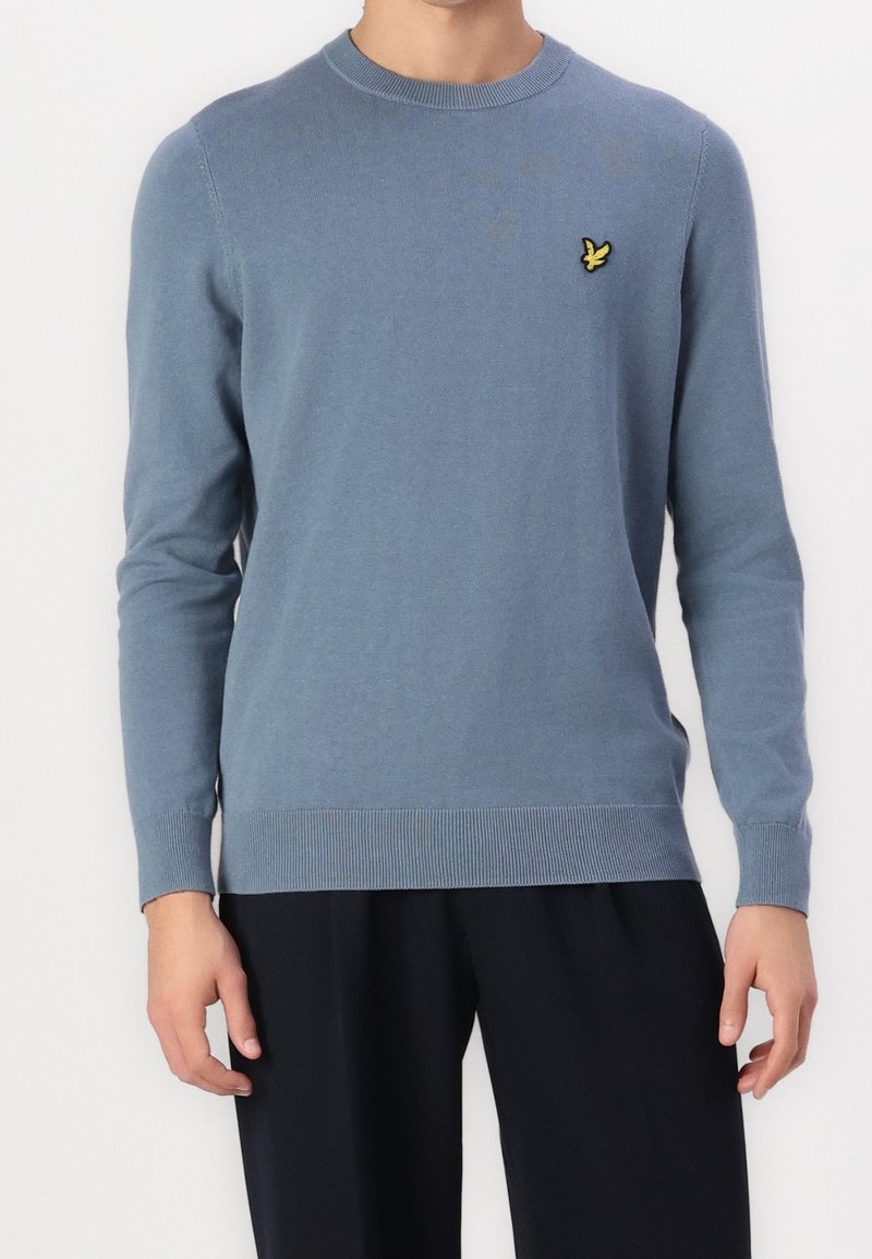 Homme portant un pull à manches longues bleu clair à col rond avec un petit logo d'oiseau jaune sur la poitrine gauche et un pantalon noir.
