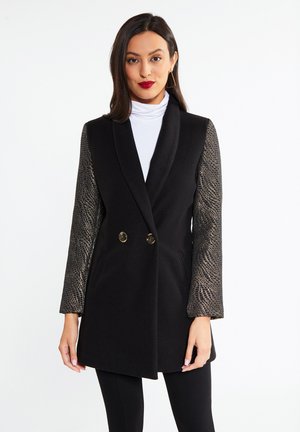 Femeie care poartă o vestă neagră fără mâneci cu mâneci lungi cu model metalic peste un guler alb, stând pe un fundal alb simplu.