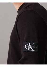 Svart texturerad långärmad tröja med rund halsringning och en svart tygpatch med "Calvin Klein Jeans" logotypen på vänster ärm.