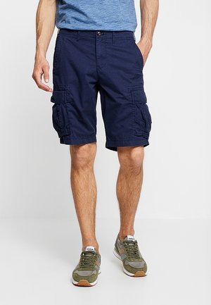 Mand iført marineblå cargoshorts, blå tekstureret t-shirt og grønne sneakers, står foran en ensfarvet baggrund.