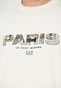 Bílý bavlněný tričko s velkým, pestrobarevným nápisem "PARIS" a městskou grafikou uvnitř, a logem "EA7 Emporio Armani" pod ním.