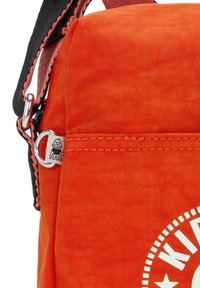 Oranžová látková taška s jednoduchým designem, černým popruhem, stříbrným kováním a bílým logem s kulatým okrajem a stylizovaným textem.