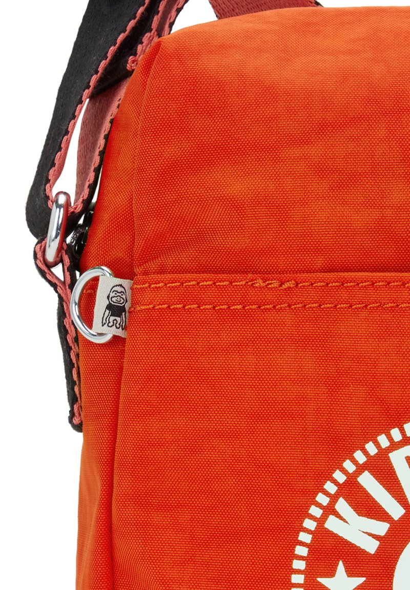Borsa in tessuto arancione con un design semplice, tracolla nera, accessori in metallo argentato e un logo bianco con un bordo rotondo e testo stilizzato.