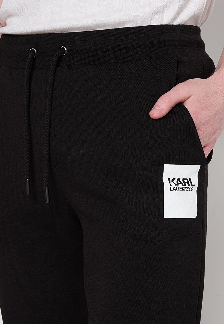 Schwarze Sweatpants aus weichem Stoff, mit einem weißen Logopatch am linken Oberschenkel, einem elastischen Bund und verstellbaren Kordeln.