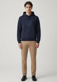 Felpa blu navy con cappuccio e coulisse, con un logo grafico rotondo sul petto. Abbinata a pantaloni color sabbia e scarpe nere.