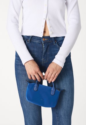Person in weißem, geripptem Langarmshirt und blauen Jeans hält eine kleine blaue Handtasche mit zwei Griffen vor sich.