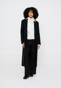 Casaco longo preto sobre um top branco com textura e calças pretas de perna larga. Modelo usa sapatos pretos e posa contra um fundo liso.