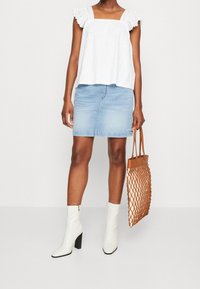 Hvid top med blonder, lyseblå denimnederdel, hvide ankelstøvler og brun flettet håndtaske, der skaber et afslappet outfit med markante teksturer.