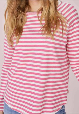 Femme portant un t-shirt à manches longues rayé horizontalement rose et blanc et un jean bleu, avec des cheveux blonds lâches tombant sur les épaules.