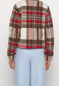 Giacca in pile a quadri con colori rosso, marrone e grigio. Caratterizzata da una vestibilità comoda, maniche lunghe e orlo corto. Abbinata a pantaloni azzurri chiari.