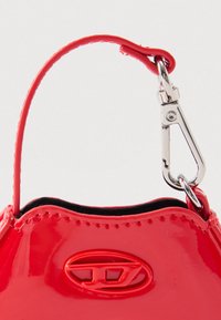 Lesklá červená rukojeť kabelky s černým lemováním, vybavená stříbrnou kovovou sponou a vyraženým logem na lesklém povrchu kabelky.