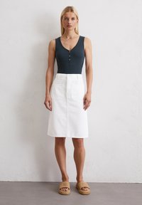 Witte denim rok met twee voorzakken, gecombineerd met een donkere tanktop met een v-hals en knoopdetails. Tan gevlochten instapschoenen.