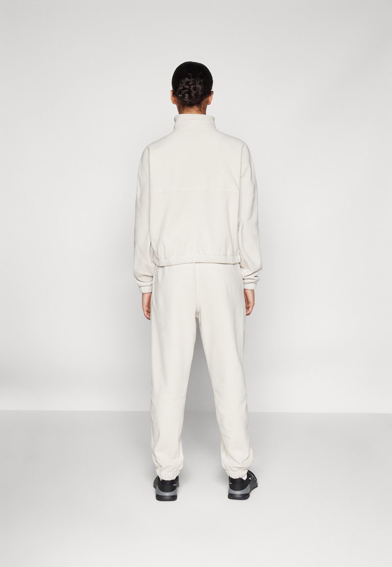 Jasnoszary, polarowy dres z wysokim kołnierzem, elastycznymi mankietami i pasem, w połączeniu z czarnymi butami. Pokazano widok z tyłu.