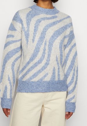 Kvinde iført en blå og hvid stribet blød strikket sweater med ribkanter på ærmer og kant, kombineret med lyse beige bukser.