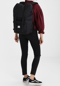 Czarny plecak z tkaniny o designie z możliwością rolowania, z zapięciami na klamry z przodu i białą naszywką z logo. Nosi go osoba w ciemnych dżinsach i sneakersach.