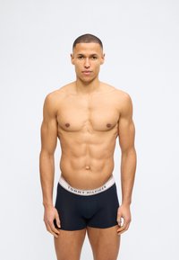 Μυώδης άνδρας με κοντά μαλλιά που φοράει μποξεράκια Tommy Hilfiger σε σκούρο μπλε, στέκεται μπροστά σε έναν απλό λευκό φόντο.