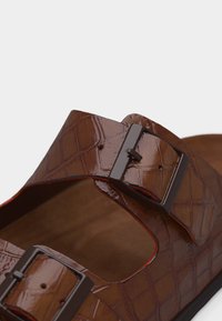 Καφέ σανδάλι τύπου slide από δερματίνη με υφή κροκοδείλου, με δύο μαύρες λουρίδες με τετράγωνες αγκράφες και επίπεδη σόλα, σε λευκό φόντο.