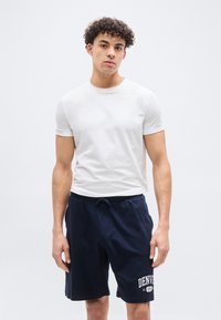 Νεαρός με σγουρά μαλλιά, που φοράει απλό λευκό μπλουζάκι και σκούρο μπλε σορτς με τη λέξη "Denver" σε λευκό φόντο.