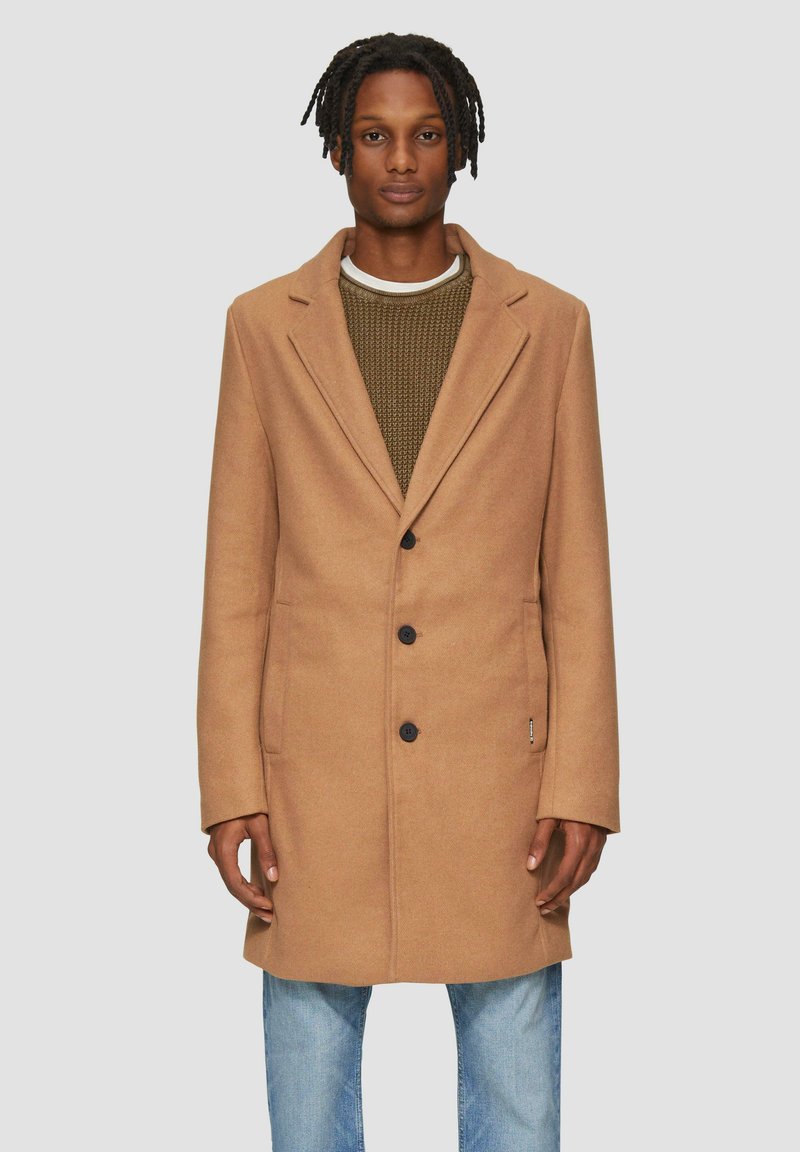 Manteau en mélange de laine brun clair avec un col cranté, quatre boutons et des poches latérales, superposé sur un pull vert texturé et un jean bleu.