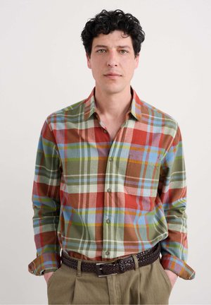 plaid button-up shirt featuring green, red, blue, and yellow tones; long sleeves; single chest pocket; worn with brown braided belt.