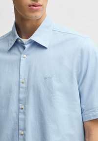 Camicia a maniche corte di colore azzurro chiaro in tessuto morbido, con colletto classico, chiusura a bottoni e logo "BOSS" ricamato sul petto.