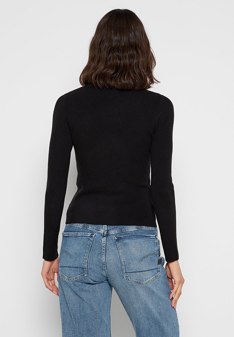 Top lungo a maniche ribbed nera con un design aderente e una texture morbida, abbinato a jeans azzurri chiari che mostrano tasche posteriori e cuciture.