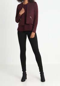 Veste bordeaux zippée à l'avant portée sur un haut assorti, associée à un pantalon noir ajusté et des bottines, mettant en valeur un design en maille texturée.