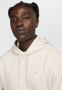 Beige Nike mīkstās auduma kapuce, ar priekšējo kangaroo kabatu, aukliņu kapuci un baltu logotipu uz krūtīm.