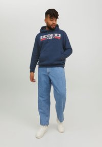 Sudadera azul marino con el logo 'JACK & JONES' en el pecho, combinada con jeans de denim azul claro y zapatillas blancas. Diseño de atuendo casual.