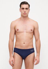 Tanga de natação azul marinho feita de um tecido suave, com um design de cintura baixa e detalhe de logotipo do lado esquerdo. Estilo minimalista com textura lisa.