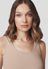 Naga top bez rękawów z okrągłym dekoltem i wąskimi ramiączkami, wykonany z gładkiego, elastycznego materiału. Modelka ma średniej długości falowane włosy.