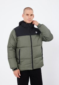 Chaqueta tipo plumón en verde oliva con sección superior negra, presenta un cuello alto, cremallera frontal y bolsillos laterales con acentos en negro.