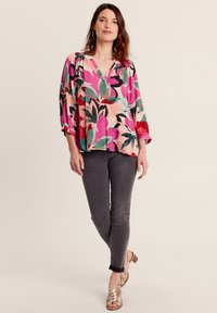 Blusa floreale in rosa, verde e nero con scollo a V e maniche a tre quarti, abbinata a jeans grigio scuro e sandali dorati.
