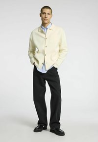 Cream button-up cardigan with a single chest pocket, layered over a light blue shirt. Paired with loose black jeans and black shoes.